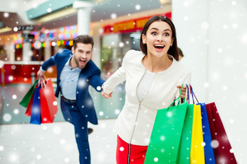 Hurry faster! It's shopping time and fun, winter sale. Handsome cheerful successful happy man holding hands and following his lovely woman with colored shopping packages, laughing in mall at holiday..