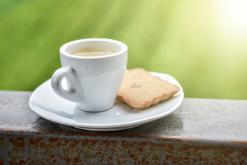 White coffee cup served with butter cookie placed on railing, view of bright morning sunlight and green garden background. Soft light effect added.
