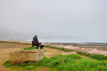 Fototapeta na wymiar Un senior qui est assis sur un banc observe un paysage brumeux sur la côte bretonne