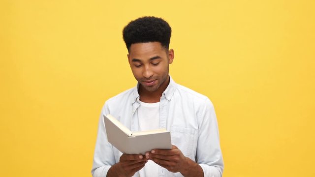 Happy african man in shirt holding and reading book over yellow background