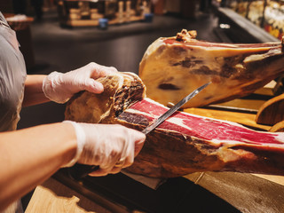Hands of a woman in gloves in the market or store cut a slice of traditionally spanish jamon de bellota. Meat close up.
