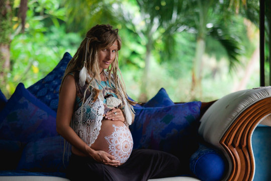 Caucasian pregnant woman with dreadlocks in boho style. White mehendi on big belly. Expectation of baby in new age lifestyle.