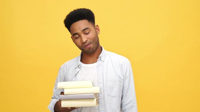 Cool Young african man in shirt holding many books and looking at the camera over yellow background