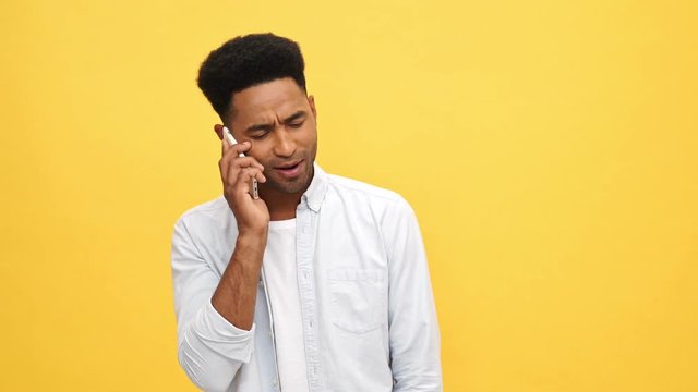 Confused Young african man in shirt talking by smartphone over yellow background