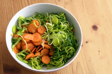 sliced fresh leek and carrots in a white bowl on a wooden table, ingredients to cook with healthy vegetables, close up