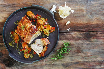Crispy roast pork with vegetables, rosemary and garlic on a rustic dark wooden table, top view from...