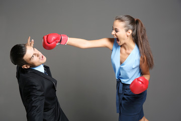 Woman in formal clothes and boxing gloves hitting businessman on grey background