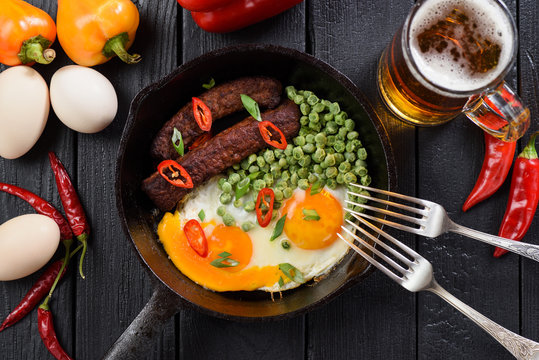 Bright protein meal of fried organic eggs, sausages and peas served for two with beer and bright chili and belle peppers on black background