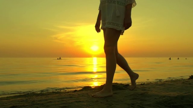 A young woman is walking along a sandy sea beach barefoot on a sunset background. The girl is walking in slow motion, the sunset is reflected on the sea.