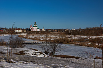 Wooden bridge over the river/City landscape of Suzdal. River, ice-bound, River banks connect a wooden bridge. In the distance, on a hill near the river stands Orthodox church.Suzdal