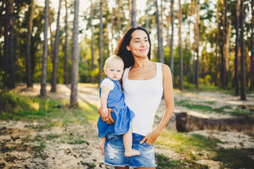 Fototapeta premium young beautiful fashionable mother with long brunette hair holding a daughter's blonde with blue eyes one year of birth in a coniferous forest in a summer park.