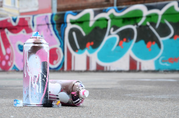 Fototapeta premium Kilka zużytych puszek natryskowych z różową i białą farbą oraz nakrętek do rozpylania farby pod ciśnieniem leży na asfalcie w pobliżu pomalowanej ściany w kolorowych rysunkach graffiti