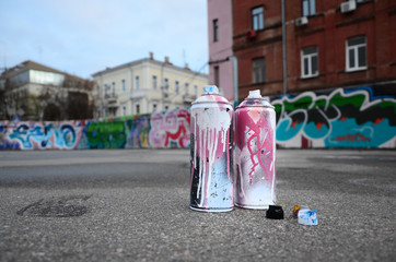Several used spray cans with pink and white paint and caps for spraying paint under pressure is lies on the asphalt near the painted wall in colored graffiti drawings