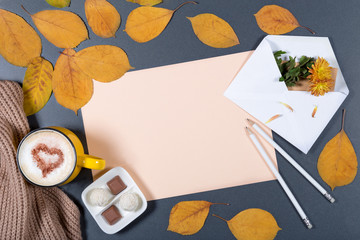 Romantic autumn mock-up. Sheet of beige paper, white envelope with note and flower, yellow and orange leaves, pencils, cup of cappuccino with heart on foam and candies.Top view.