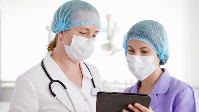 Two pretty female doctor colleagues in medical masks and caps discussing something on tablet at modern hospital room. Medical professional staff at work