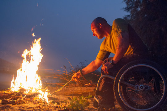 A paraplegic man sitting in a wheelchair stokes a campfire on a beach at night.