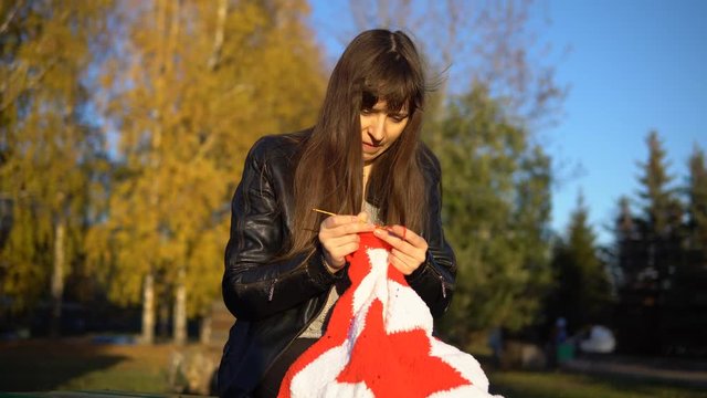 A young brunette woman in a black leather jacket crochets knit a red and white plaid in the shape of a star, sitting in an autumn or winter park. A girl makes a gift by hand.