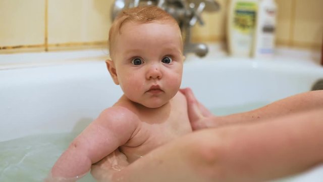 cute gentle toddler baby boy bathing white tub bathroom mother hands helping water surprised eyes nice face close up portrait looking camera calm child sweet chubby cheeks mom motherhood health caring