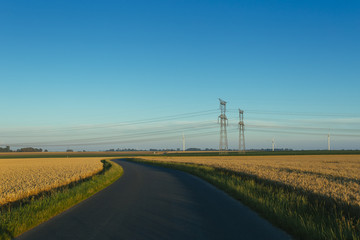 High voltage power lines and transmission towers. Asphalt road passing through agricultural fields in Normandy, France. Electric power industry, road network and agriculture concept