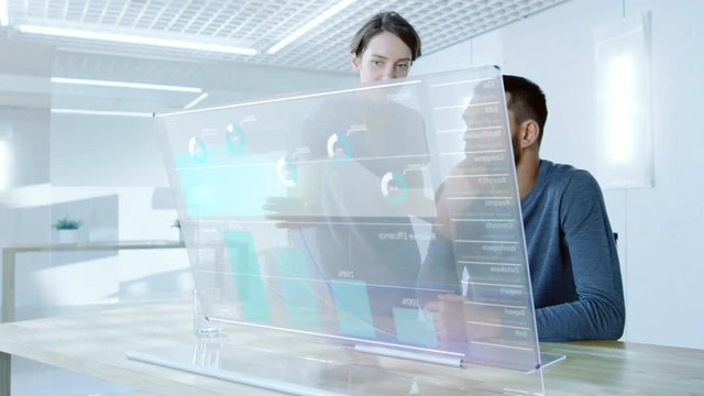 In the Near Future Male and Female Office Workers Discuss Graphical Statistic Shown on Transparent Computer Display. Beautiful People Talking. Interactive Charts on the Screen. 