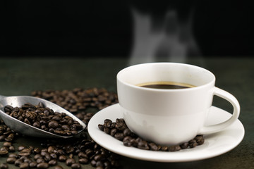 Hot coffee in the white cup with roast coffee beans and scoop on stone table in black background
