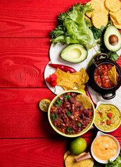 Vegetarian Mexican food concept: refried black and red beans. guacamole, salsa, chili, tortilla chips and fresh ingredients over vintage red rustic wooden background. Top view, flat lay