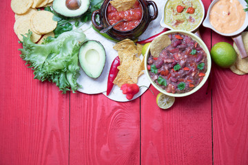 Fototapeta na wymiar Vegetarian Mexican food concept: refried black and red beans. guacamole, salsa, chili, tortilla chips and fresh ingredients over vintage red rustic wooden background. Top view, flat lay