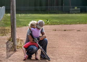 An uncle wearing a hat and baseball gloves kneeling down and giving his neice a comforting hug at a...