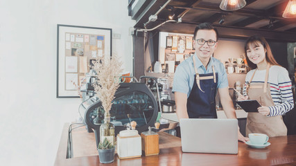 Young Asian man&woman barista wear apron using laptop with coffee cup served to customer at bar counter in coffee shop with smile face.Concept of cafe and coffee shop small business.Vintage tone