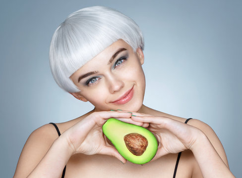 Pretty girl with green avocado. Photo of blonde girl holding a half of avocado on blue background. Skin care and beauty concept.