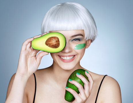 Happy girl with green avocado. Photo of blonde girl holding a half of avocado in front of her face and smiling on blue background. Skin care and beauty concept.