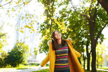 A beautiful happy cheerful brown-haired woman in a yellow coat and striped longsleeve rejoices with a tablet in her hands and white headphones in fall city park on a warm day. Autumn golden leaves.