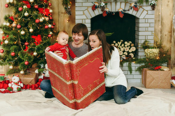 Fototapeta na wymiar Young playful happy parents with a cute little baby boy reading big red book in a decorated room at home. Christmas good mood. New Year. Lifestyle, family and togetherness concept.