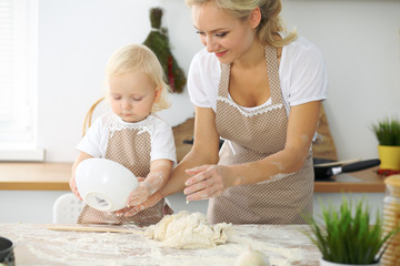 Obraz na płótnie Canvas Little girl and her blonde mom in red aprons playing and laughing while kneading the dough in the kitchen. Homemade pastry for bread, pizza or bake cookies