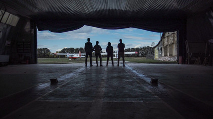 Four people standing in an aircraft hangar silhouetted against the sky watching two small aircraft outside in a low angle view - obrazy, fototapety, plakaty