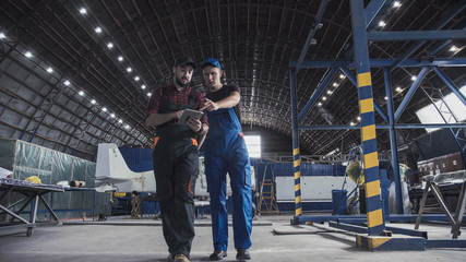 Two flight engineers walking through a large aircraft hangar talking and gesturing together - obrazy, fototapety, plakaty