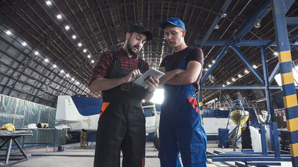 Two flight engineers walking through a large aircraft hangar talking and gesturing together