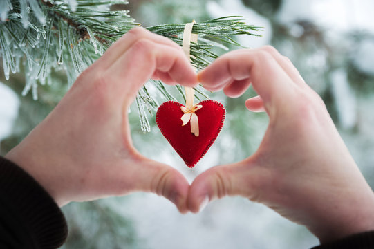 Male hands in the shape of a heart on a background of toys made of red felt. Beautiful snow-white white forest in winter. Background for your text from snow-covered branches and paws of Christmas tree