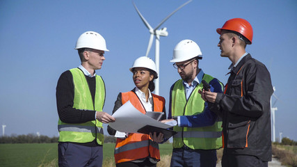 Four mixed ethnic people engineers and workers in multicolored jackets and hard hats standing in...
