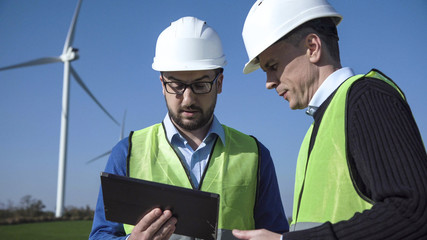 Two engineers wearing hard hat discussing over digital table against turbines on wind farm