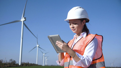 Caucasian female engineer wearing hard hat standing with digital tablet against wind turbine on...