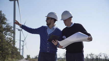 Two engineers discussing a blueprint on a wind farm in front of a turbine supplying alternative electricity from the conversion of kinetic energy