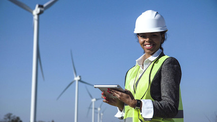Female engineer wearing hard hat standing with digital tablet against wind turbine on sunny day and...