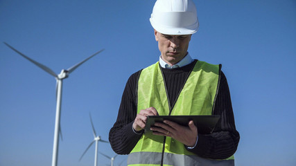 Engineer with seriously face working with digital tablet against wind turbine on sunny day and looking at camera © Framestock