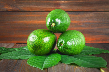 Three green raw ripe avocados with leaves lie on a wooden brown table.