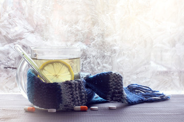 treatment of colds in winter/ hot infusion with lemon in mug, wrapped in a scarf with a thermometer on the background of pills and frozen windows