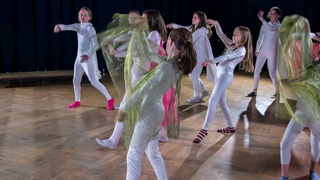 Girls in tight white costumes and green capes are dancing on stage and are performing in a school play.