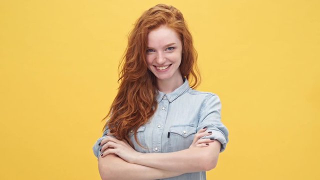 Happy winner ginger woman in denim shirt holding crossed arms and looking at the camera over yellow background
