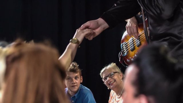 Music band performing on the stage in front of their fans and atractive male singer touches hands with female fan.