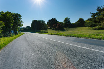 Empty asphalt country road passing through green fields and forest. Countryside landscape on sunny day with sunbeams in the blue sky in France. Transport, industrial agriculture, road network concept.
